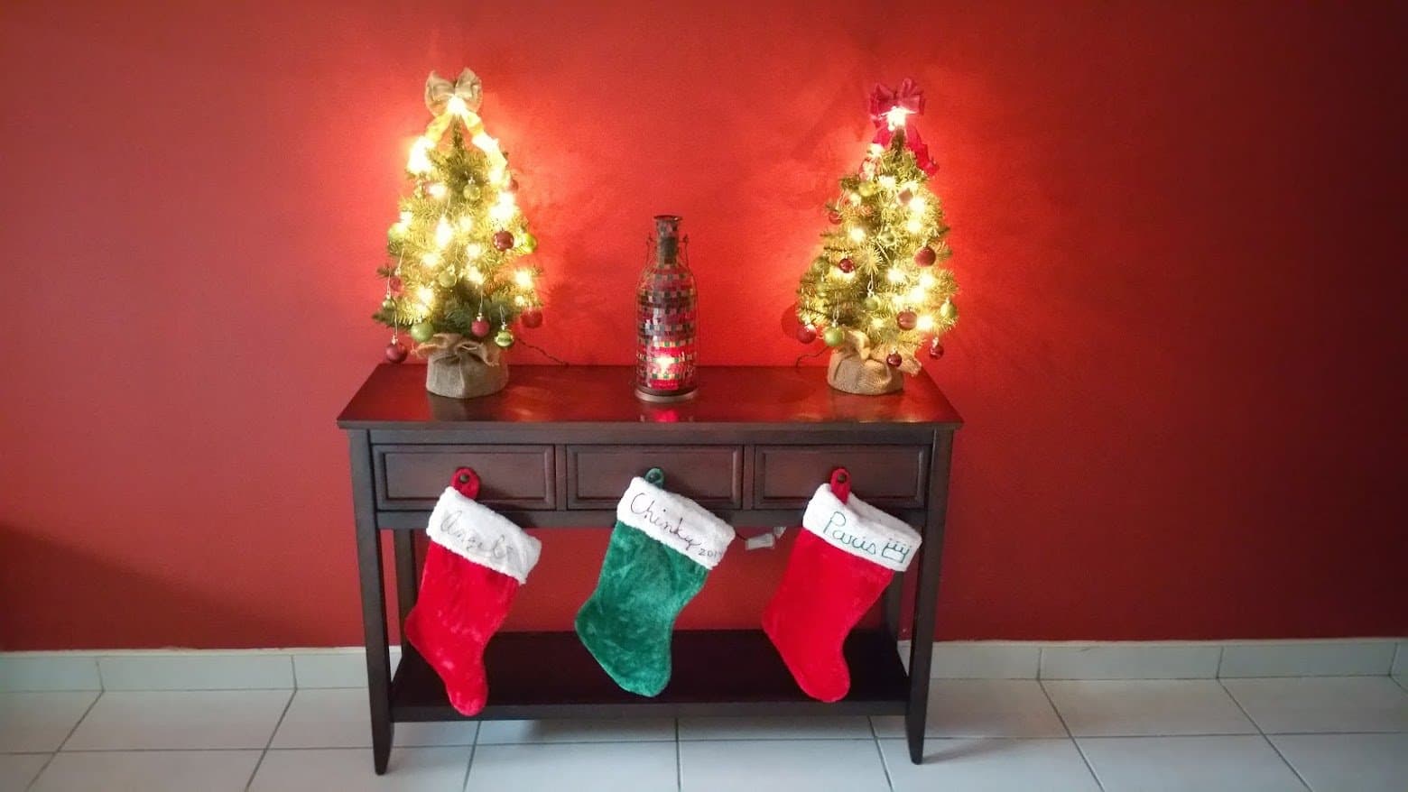 Two small lit Christmas trees on a table with three named family stockings hanging below